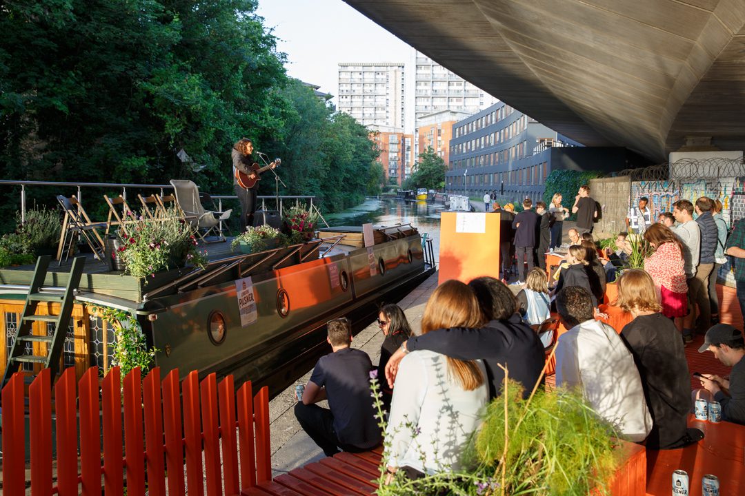 A group of people sitting on the side of the canal and a performer playing guitar on a boat