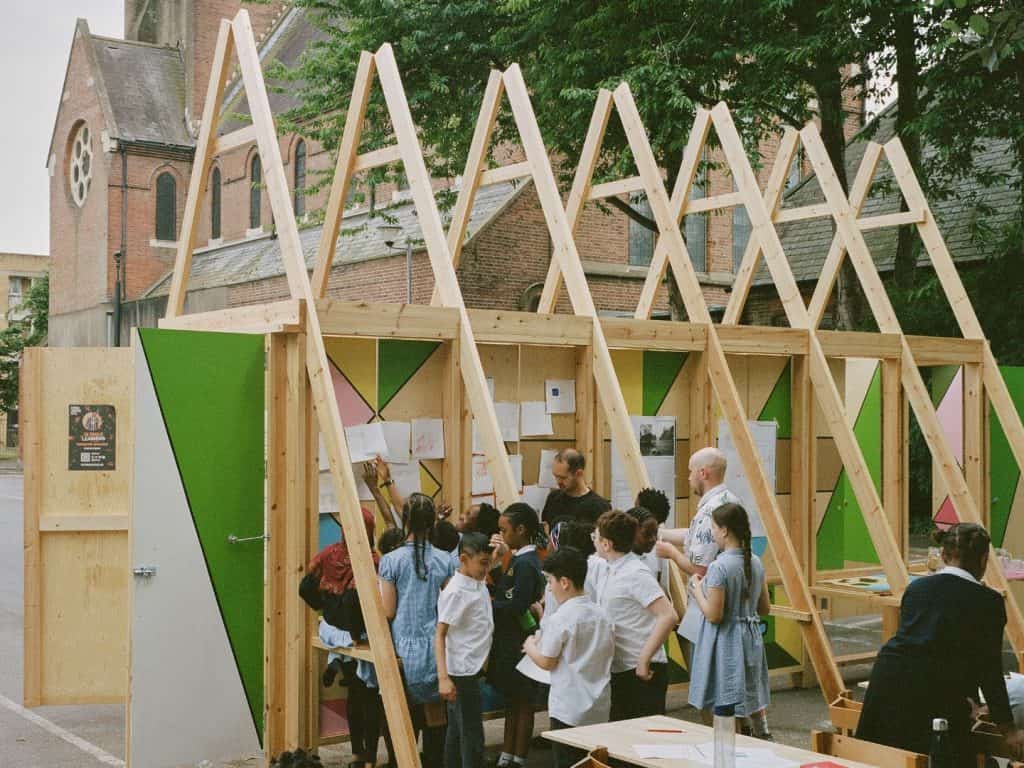 Children stand next to a wooden structure with drawings pinned on.